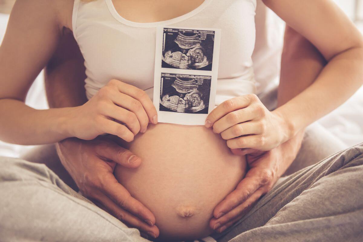 Na zdjęciu widoczny jest ciążowy brzuch kobiety, na którym dłonie ułożone są w kształt serca. Delikatne światło oraz jasne, rozmyte tło tworzą spokojną i ciepłą atmosferę, kojarzącą się z oczekiwaniem na narodziny dziecka. Fotografia symbolizuje macierzyństwo, troskę o zdrowie matki i dziecka oraz przypomina, jak ważna jest dbałość o dobrostan psychiczny kobiety w okresie ciąży i po porodzie. Nawiązuje do tematyki depresji poporodowej oraz znaczenia wczesnego rozpoznania objawów i odpowiedniego wsparcia emocjonalnego.